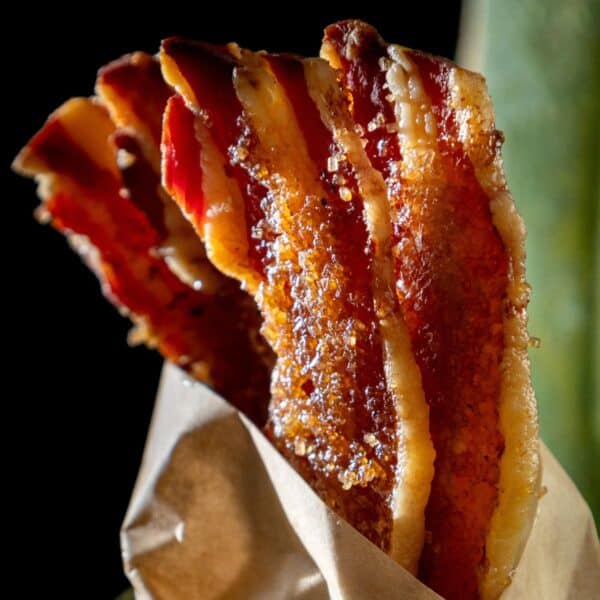 Three strips of candied bacon wrapped in brown parchment paper with a dark background.