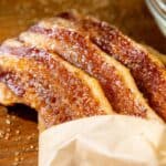 Four strips of candied bacon coated with brown sugar, partially wrapped in parchment paper on a wooden surface.