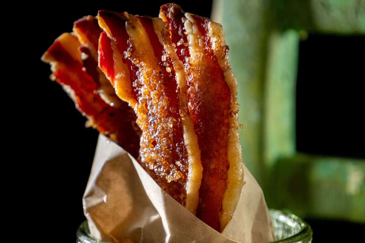 Strips of candied bacon stand upright in a brown paper wrapper on a dark background.