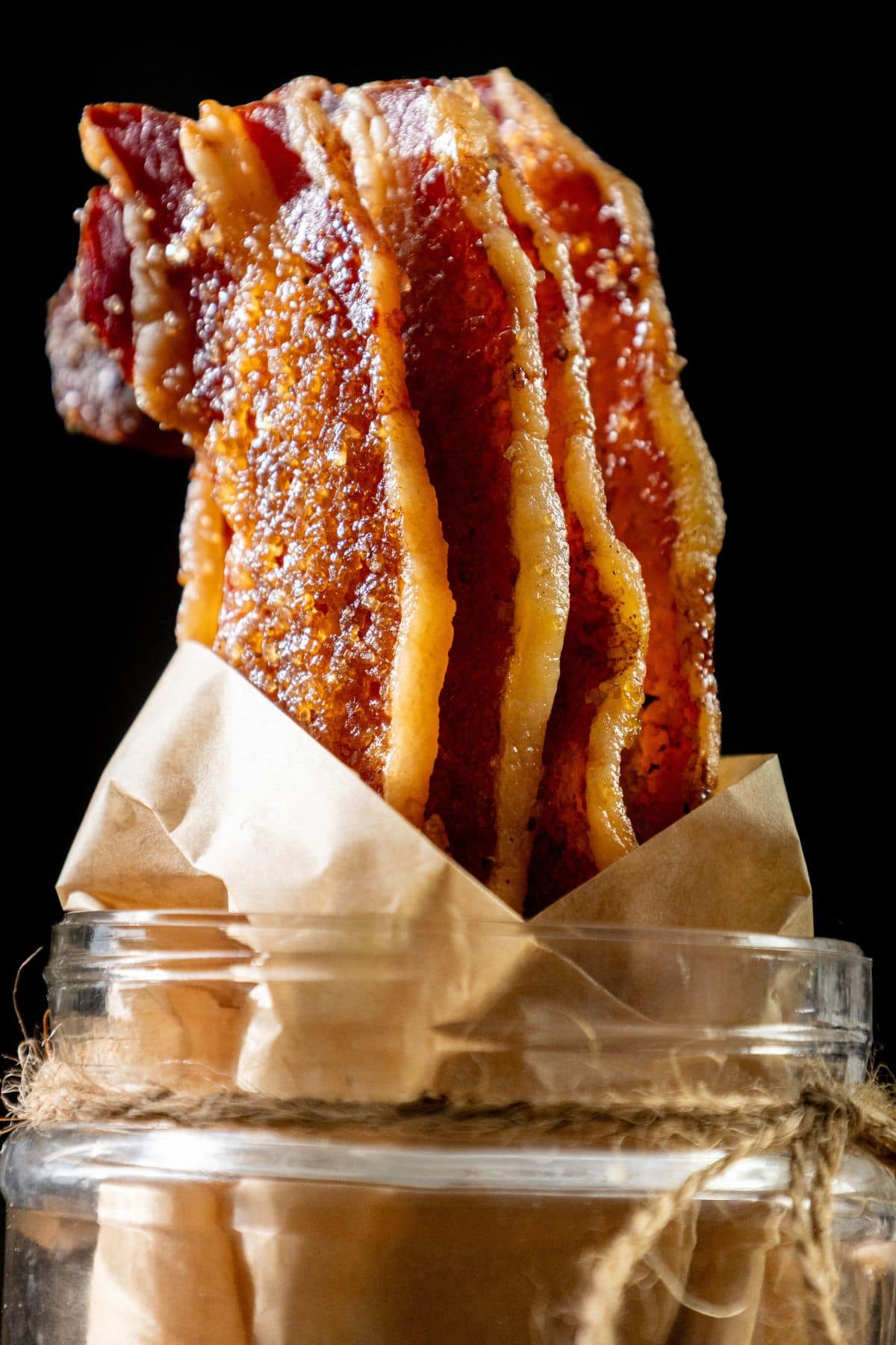 Candied bacon strips stand upright in a glass jar lined with brown parchment paper and tied with twine on a dark background.