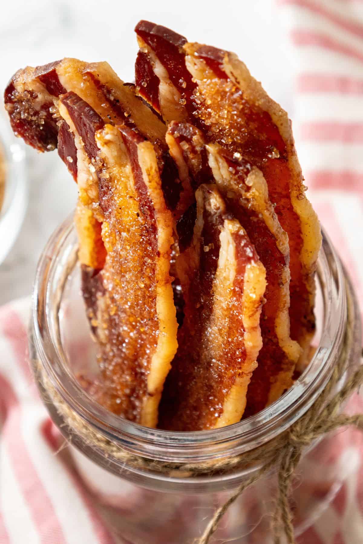 Strips of candied bacon stand upright in a glass jar with a red and white striped cloth in the background.