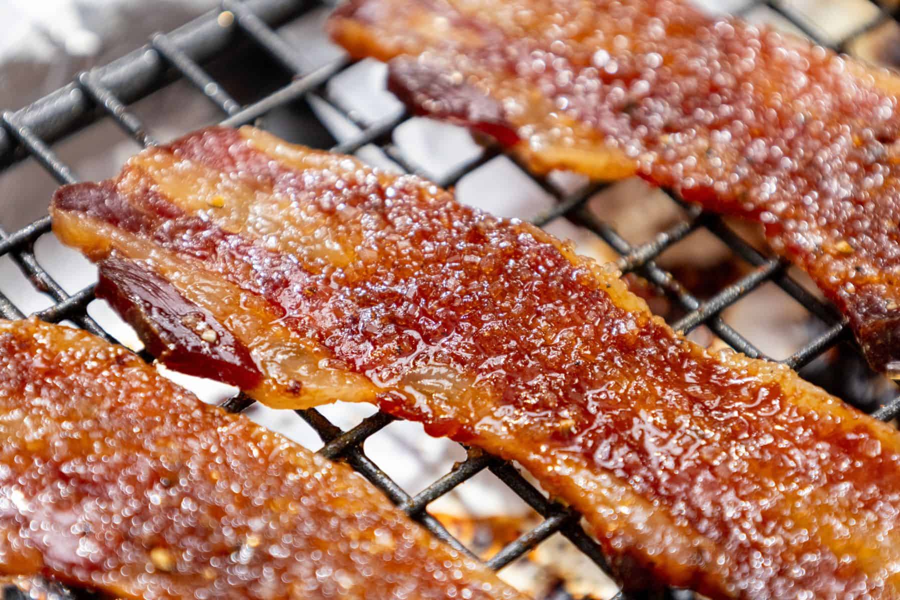 Several strips of candied bacon on a metal grill, coated with seasoning and glistening rendered fat.