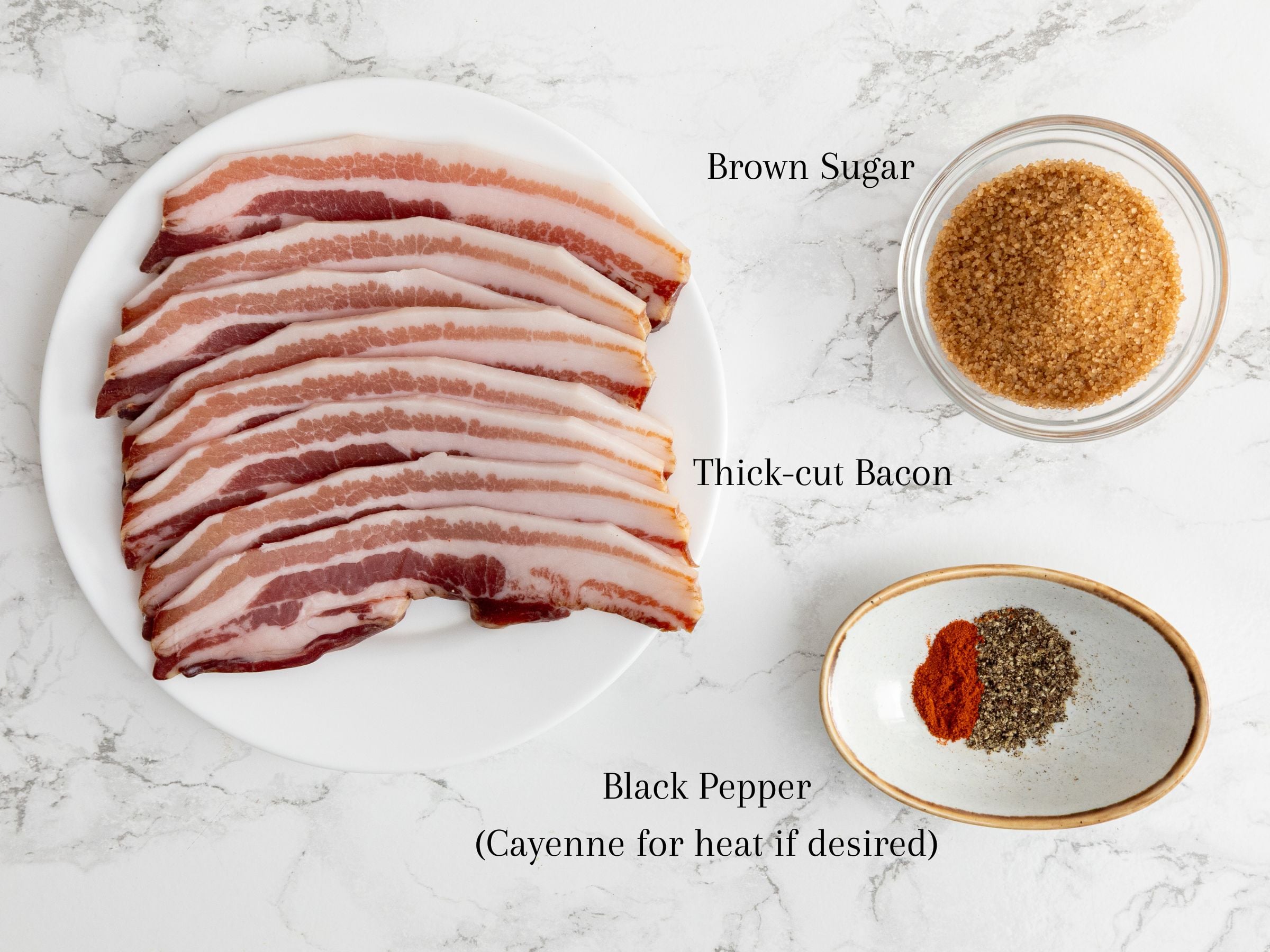 Oven baked candied bacon slices on a plate, a bowl of brown sugar, and a dish with black pepper and cayenne pepper on marble.