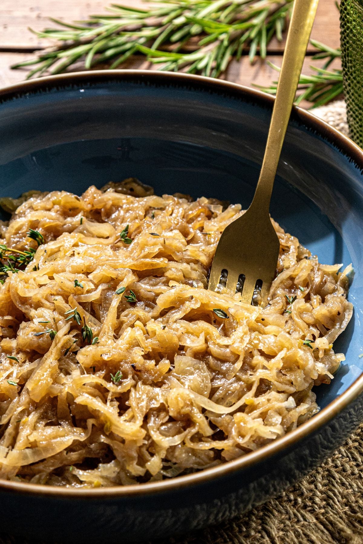 A bowl of caramelized onions with herb garnish, a fork inside, and rosemary sprigs in the background.