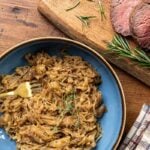 Caramelized onions with herbs on a blue plate beside sliced roast beef and rosemary on a wooden board.