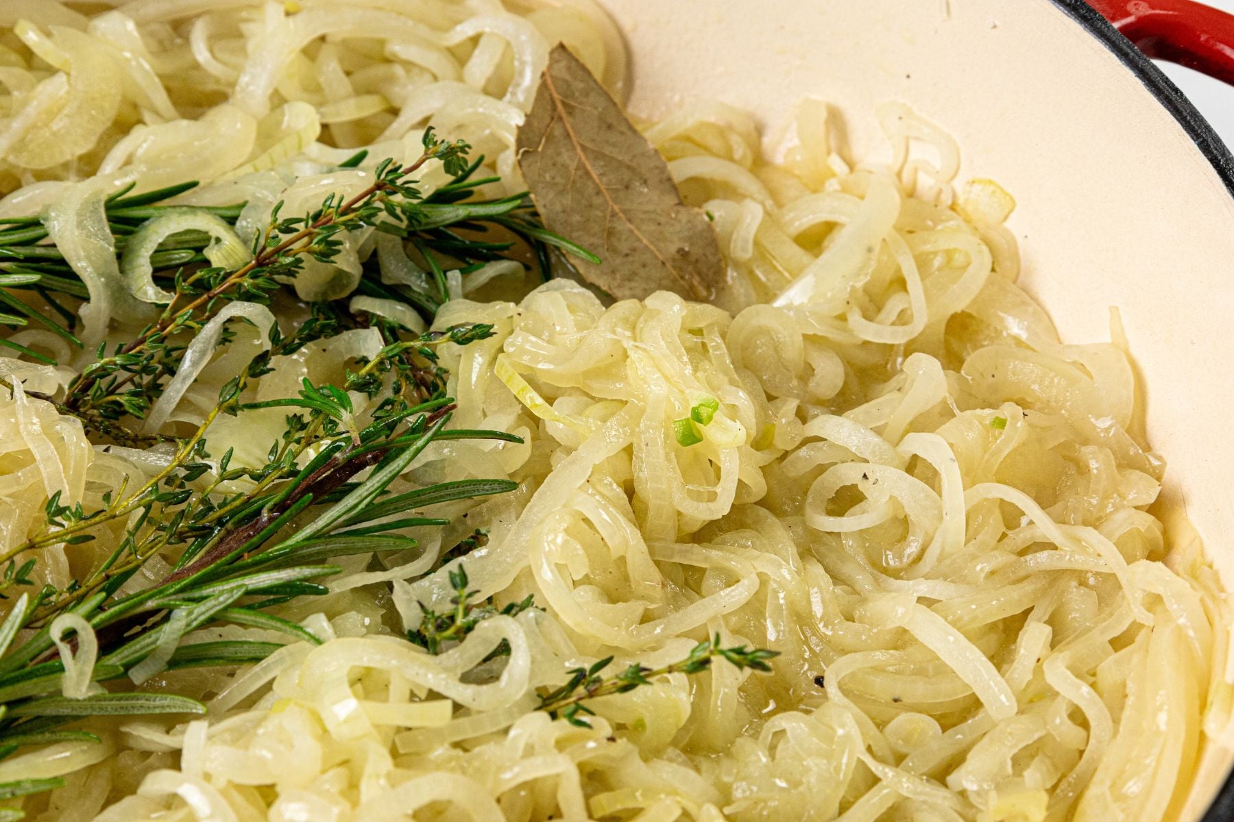 Sliced onions in a pan with sprigs of rosemary, thyme, and a bay leaf.