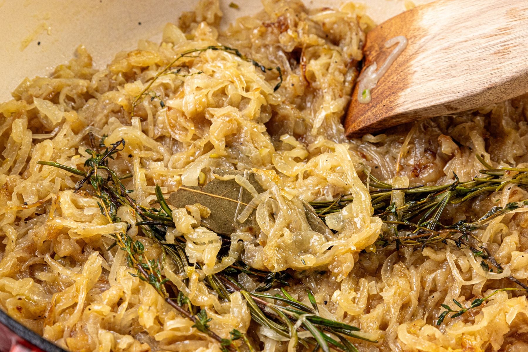 Caramelized onions in a pan with fresh thyme and a bay leaf, being stirred with a wooden spoon.