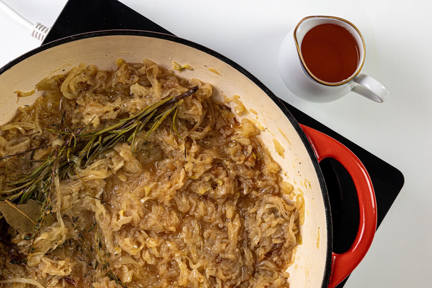 A red Dutch oven with caramelized onions and herbs beside a small white pitcher of broth on a clean white surface.